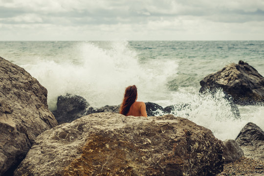 A naked young girl on the ocean coast during a storm covered herself with a stone on the beach. The surging, mighty, strong waves. Storm in the ocean. Beach and stones.Storm on the sea. Gray and blue.