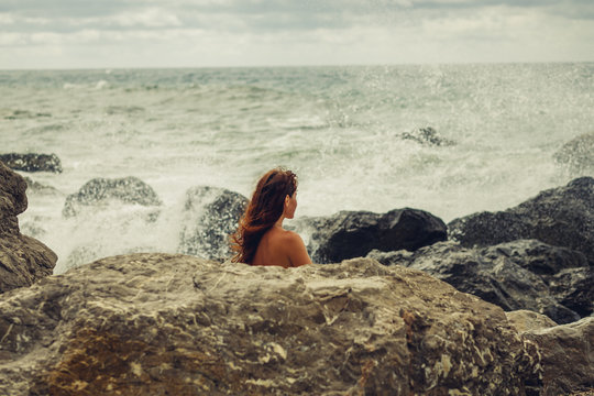 A naked young girl on the ocean coast during a storm covered herself with a stone on the beach. The surging, mighty, strong waves. Storm in the ocean. Beach and stones.Storm on the sea. Gray and blue.