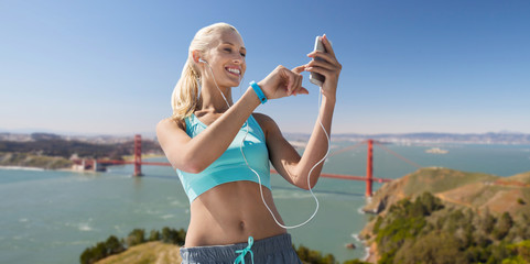 sport, technology and healthy lifestyle concept - smiling young woman with smartphone, earphones and fitness tracker listening to music over golden gate bridge in san francisco bay background