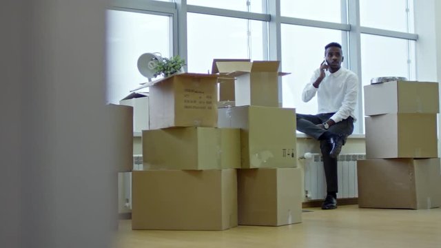 Tracking shot of african american businessman sitting on window sill in new office with stacks of cardboard boxes and talking on cell phone