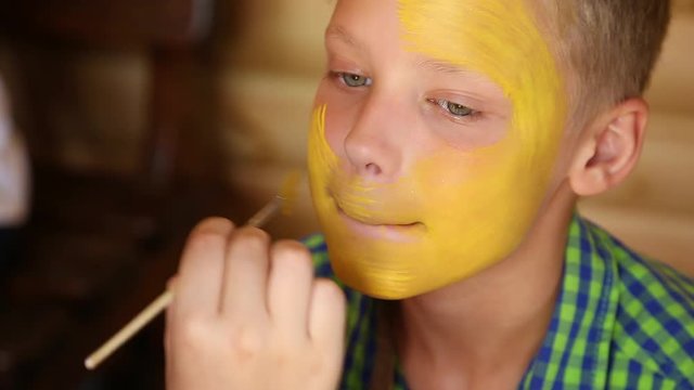 Woman paints face of cute kid with yellow and white facepaints at birthday party.