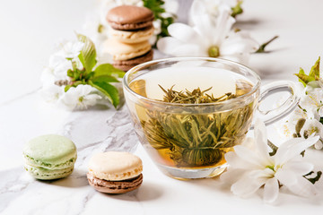 Glass cup of hot green tea with french dessert macaroons, spring flowers white magnolia and cherry blooming branches over white marble texture background.