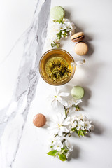Fototapeta na wymiar Glass cup of hot green tea with french dessert macaroons, spring flowers white magnolia and cherry blooming branches over white marble texture background. Top view, copy space.