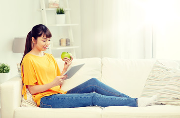 people, healthy eating, education, technology and concept - happy young asian woman sitting on sofa with tablet pc computer and green apple at home