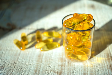 Yellow gold oil vitamin, omega 3 capsules in a glass beaker in the rays of light on a wooden background. Close up, selective focus