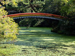 Fototapeta premium Sunlit Japanese garden with green pond and red arching bridge at Usa shrine in Oita prefecture