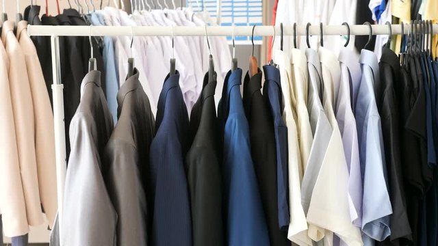 Different male business clothes, including suits and shirts on a hanger. Dolly footage. There are other clothes on hangers in the background