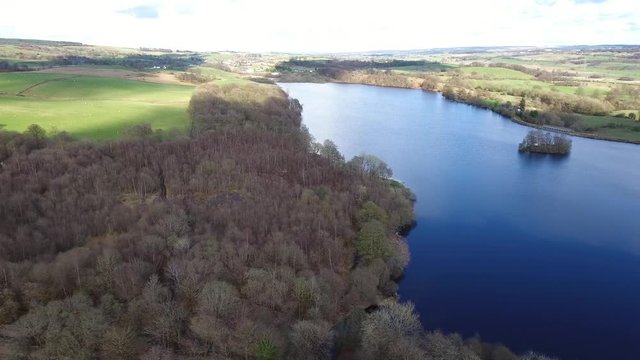 Aerial footage flying right, over Banton Loch near Kilsyth in Scotland. Panorama of the loch, little island and woodland with bright blue sky reflected on the surface.