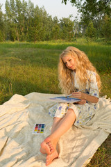 Beautiful charming barefoot long curly blonde hair teenage girl wearing a long light dress outdoors on a picnic paints with watercolor drawing sitting on a blanket on the grass.