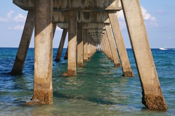 Underside of Deerfield Beach, Florida Pier with the Sun Revealing Concrete Supports and Water Lapping Underneath in Late Afternoon with a Boat Passing in the Distance