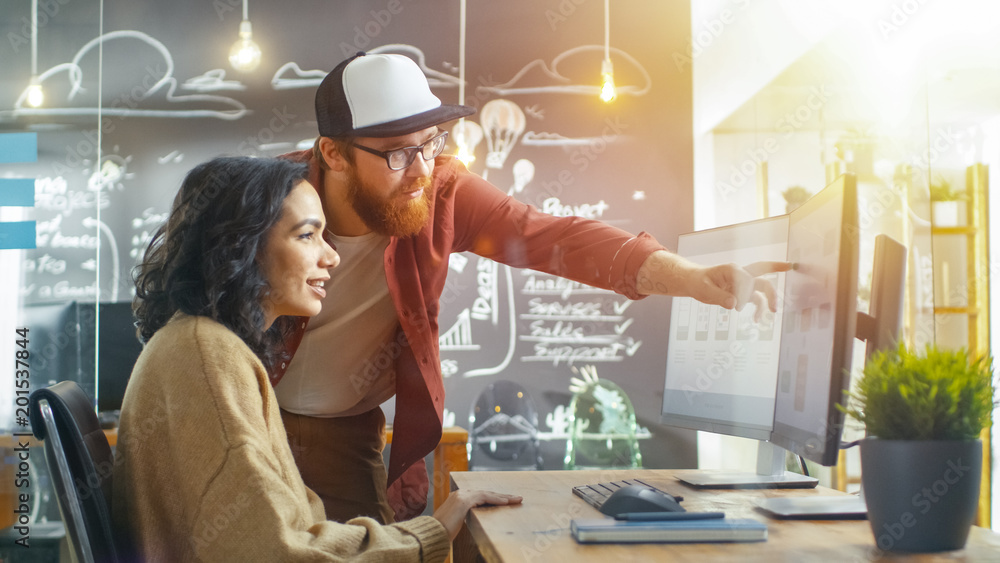 Poster at the desk beautiful man programmer helping young woman developer, pointing at the monitor, togethe