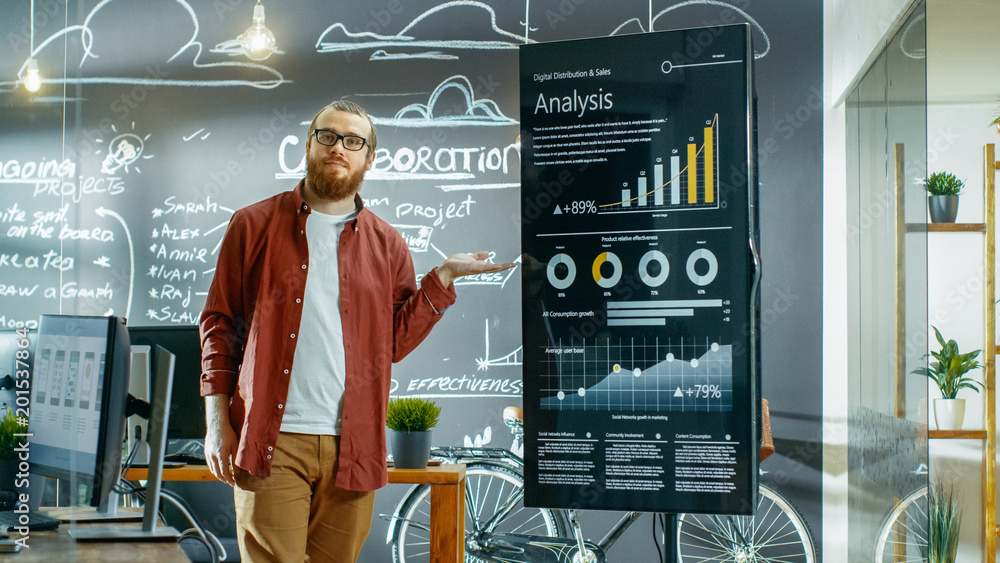 Poster Male Developer Looks at Interactive Whiteboard with Charts, Graphs and Growth Statistics. He Points at Display with Presenting Gesture and Smile. In the Background Creative Office with Chalkboard Wall