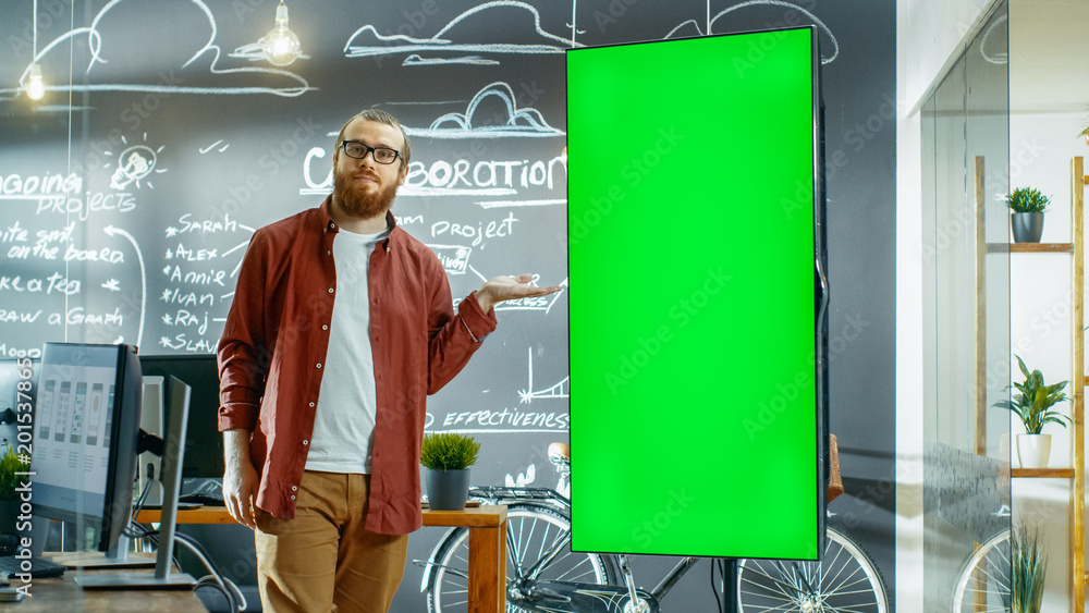 Poster Male Developer Looks at Interactive Whiteboard with Mock-up Green Screen. He Points at the Screen with Presenting Gesture and a Smile. In the Background Creative Office with Chalkboard Wall.