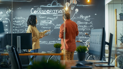 Female Project Manager Holds Tablet Computer and Shows Plan Drawn on a Blackboard Wall to Male...