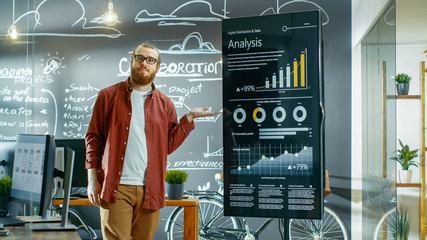 Male Developer Looks at Interactive Whiteboard with Charts, Graphs and Growth Statistics. He Points at Display with Presenting Gesture and Smile. In the Background Creative Office with Chalkboard Wall