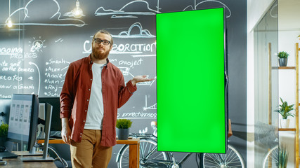 Male Developer Looks at Interactive Whiteboard with Mock-up Green Screen. He Points at the Screen with Presenting Gesture and a Smile. In the Background Creative Office with Chalkboard Wall.