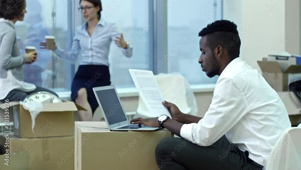 Poster tracking shot of african man typing on laptop on stack of boxes and reading document while two busin