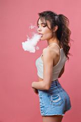 Casual pretty woman standing and vaping on pink background in studio.