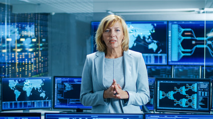 In the System Control Room Female Chief Engineer Describes Her Project Talking into the Camera. In the Background Multiple Screens Showing Interactive Data.