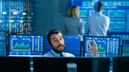 Stock Exchange Trader Makes a Deal with Big Client Over the Headset. He Works with Other Brokers and is Surround By Computers with Graphs and Ticker Numbers on Screens.