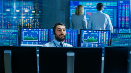 Stock Exchange Trader Makes a Deal with Big Client Over the Headset. He Works with Other Brokers and is Surround By Computers with Graphs and Ticker Numbers on Screens.