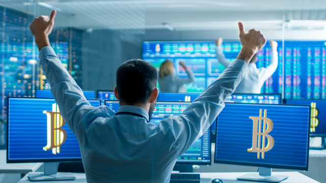 Back View of the Stock Trader Raising His Arms in Big Victory, He's Working for the Cryptocurrency Exchange Department. Screens Show Relevant Data, Graphs and Ticker Numbers.