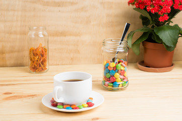 Coffee mug, red flowers, colored candy and orange peel in a jar and a yellow diary on a wooden background. breakfast or lunch