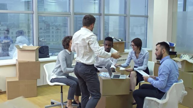 Tracking shot of diverse business team sitting in new office at stacks of boxes, discussing documents and then standing up, throwing paper and raising arms while celebrating relocation