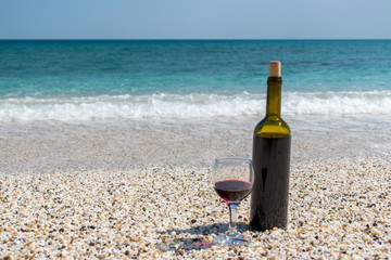 Glass of red wine and bottle on the beach at the summer sunny day. Sea on the background.