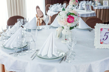 Glasses, flowers, fork, knife, napkin folded in a pyramid, served for dinner in restaurant with cozy interior. Wedding decorations and items for food, arranged by the catering service on a large table