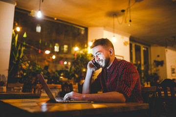 Young handsome Caucasian man with beard and toothy smile in red shirt works behind laptop, hands on keyboard sitting at wooden table. Uses calls on mobile phone. In evening at the coffee shop