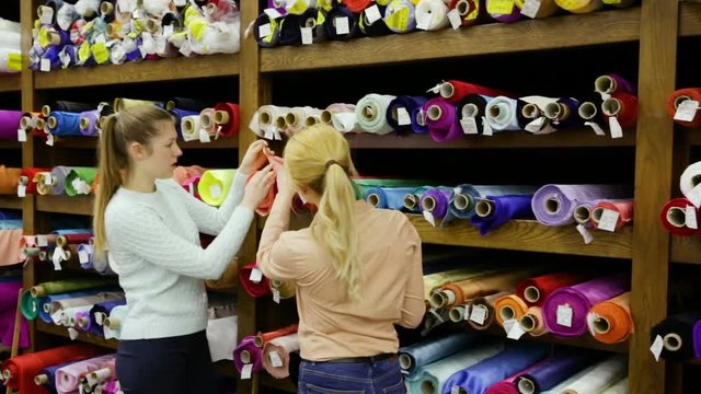 Two cheerful young female friends looking for interesting cloth in textile shop