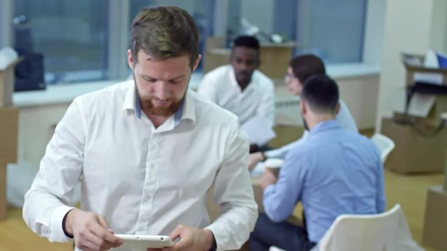 Young bearded businessman using digital tablet and posing at camera in new office after moving in while his colleagues having meeting in background