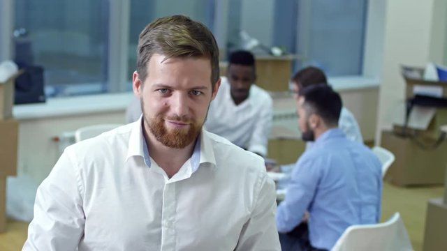 Portrait of young bearded businessman using digital tablet and smiling at camera in new office after relocation while his colleagues having meeting in background