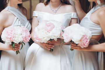 beautiful bridesmaids and bride holding stylish peony bouquets in the morning in hotel room....