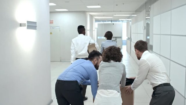 Rear view of colleagues walking with belongings and team of office workers pushing large heavy box through hallway of new modern office building