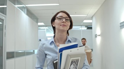 Young attractive businesswoman walking through hallway in new office center with personal belongings and smiling while looking at excited colleagues running with cardboard boxes - Powered by Adobe
