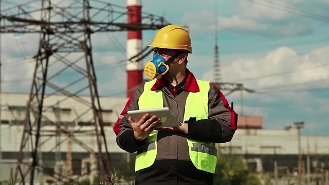 Powerman in gas mask with tablet computer on electric power station. Power engineering specialist in respirator and yellow hard hat with personal computer in hands at heat power plant