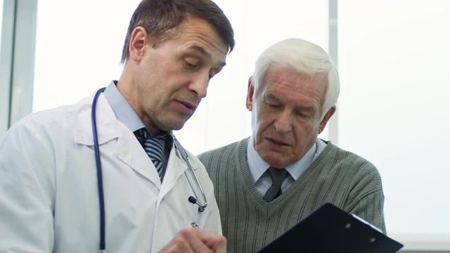 Medium shot of mature physician with clipboard showing test results to senior patient and explaining something
