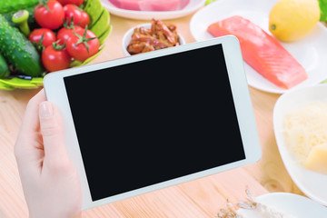 Close-up top view of female hands holding a tablet computer with a blank display surrounded by healthy food on a home kitchen table. Workpiece to insert the design of the mobile application into the