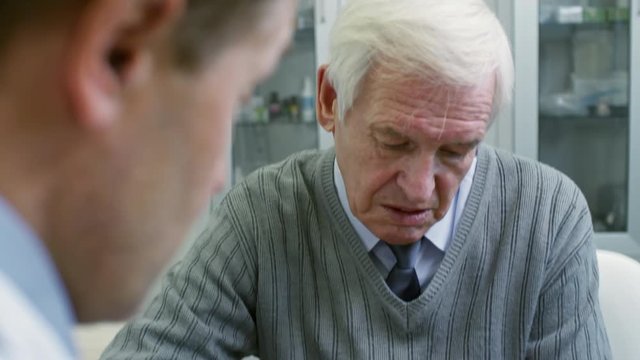 Tilt down of male physician in lab coat holding tablet and showing document to senior patient