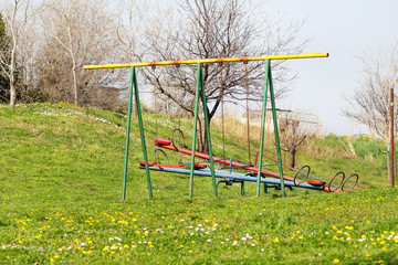 Two empty children swings and seesaw at playground activities in public park / Beautiful natural environment, grass, spring flowers / Colorful playground equipment for children / Sunny day in spring.
