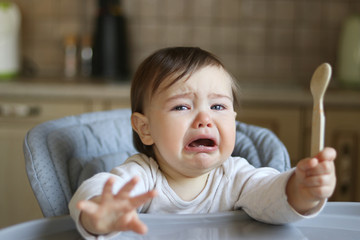 Crying hungry little baby with tears in eyes sitting in the high feeding chair at kitchen holding...