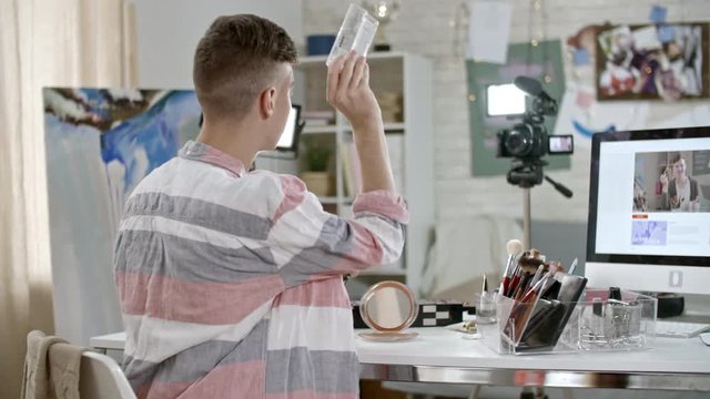 PAN of teenage boy showing eyeshadow palette to camera with led light standing on tripod and making swatches while reviewing product for video blog