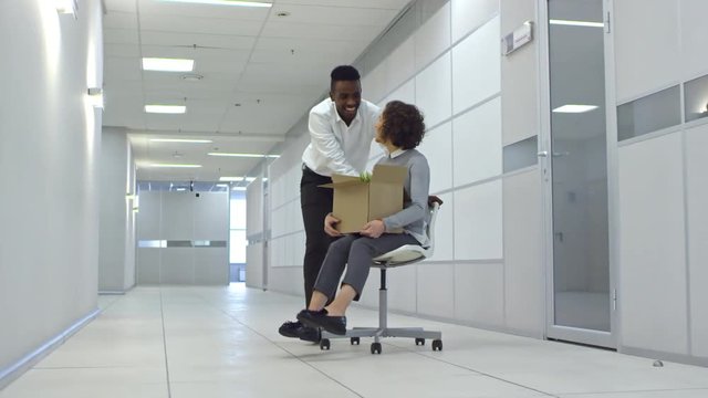 Young african american office worker running through hallway in new office building and pushing chair with female colleague towards the camera; woman holding cardboard box with belongings