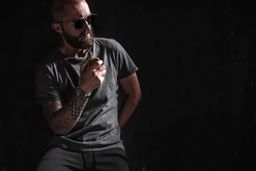 Men with beard in sunglasses vaping and releases a cloud of vapor on dark moody studio background.