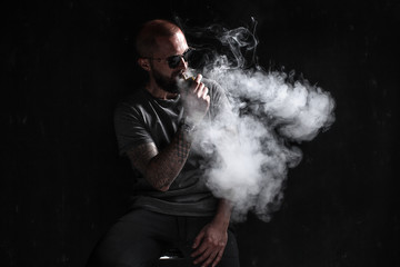 Men with beard in sunglasses vaping and releases a cloud of vapor on dark moody studio background.