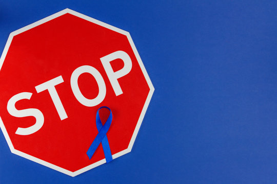 World cancer day concept. Blue ribbon as a symbol of bowel cancer and sign stop isolated on dark blue background.