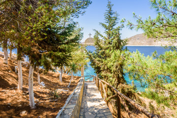 Fototapeta na wymiar Beautiful sunny coast view to the greek blue sea with crystal clear water beach near a small chapel with a white arch with holy cross situated in a small forest, Leros, Dodecanese Islands/ Greece