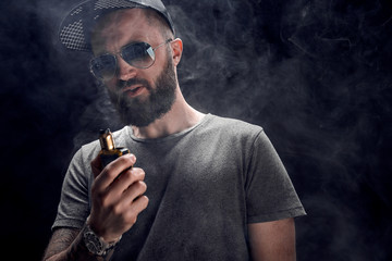 Pensive Black bearded male dressed in a grey shirt, sunglasses and baseball cap vaping. man in...